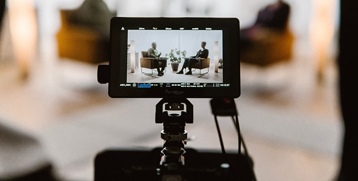 取材風景の撮影写真（椅子に座った二人が向かい合って対談している風景をカメラで撮影しようとしている状況で、カメラのモニター画面が写真中央にきている取材風景の画像）